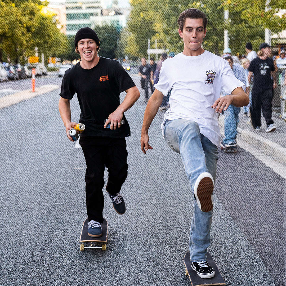 Two young men skate down a city street at dusk, one wearing a black t-shirt and black pants holding a spray can, and the other wearing a white t-shirt and light blue jeans.