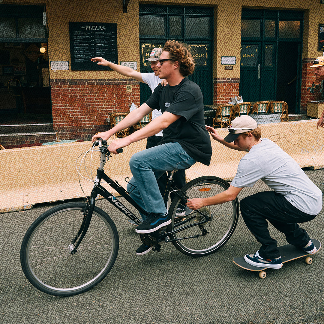 A man rides a black bicycle while another man on a skateboard pushes him from behind, with a third person pointing in the background, set against a street scene in front of a brick building.