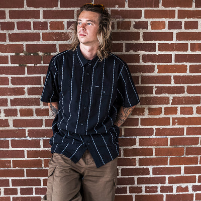 A person with long, wavy hair stands against a red brick wall, wearing a black short-sleeve, button-up shirt with white stripes and khaki pants. They look off to the side with a relaxed expression.