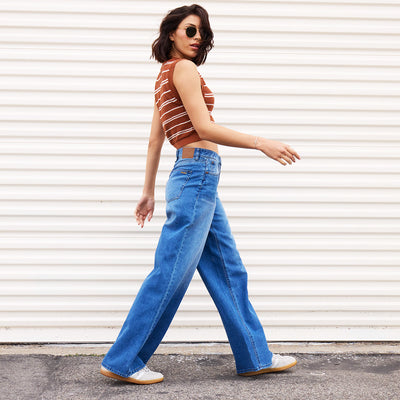 A woman wearing a striped sleeveless top, wide-leg blue jeans, white sneakers, and sunglasses walks confidently in front of a white corrugated wall.