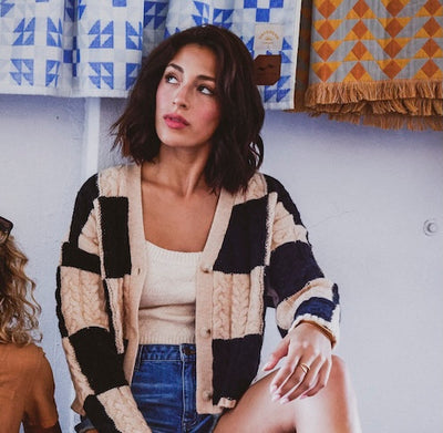 A woman wearing a patchwork knit cardigan with beige, black, and navy blue panels over a cream tank top and blue jeans, seated against a white wall with patterned textiles in the background.