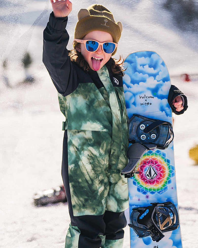 Child in green snowsuit holding a blue and white snowboard with a colorful design, standing in snow.