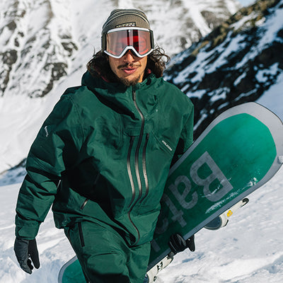 Person in green snowboarding gear holding a green snowboard with 'Batac' branding in a snowy mountain setting.