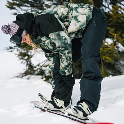 Person adjusting ski boots in a snowy landscape with trees in the background