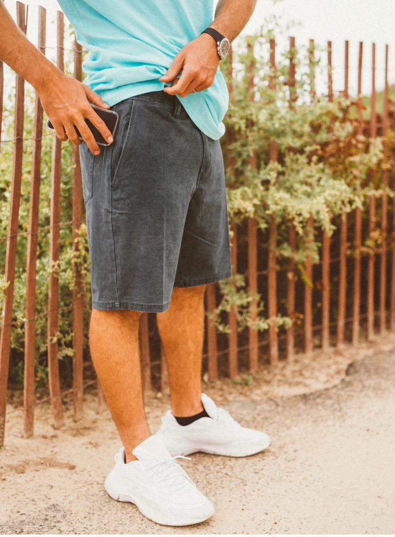 A person wearing a turquoise shirt, dark gray shorts, and white sneakers stands outdoors near a wooden fence and green bushes, holding a phone in one hand.