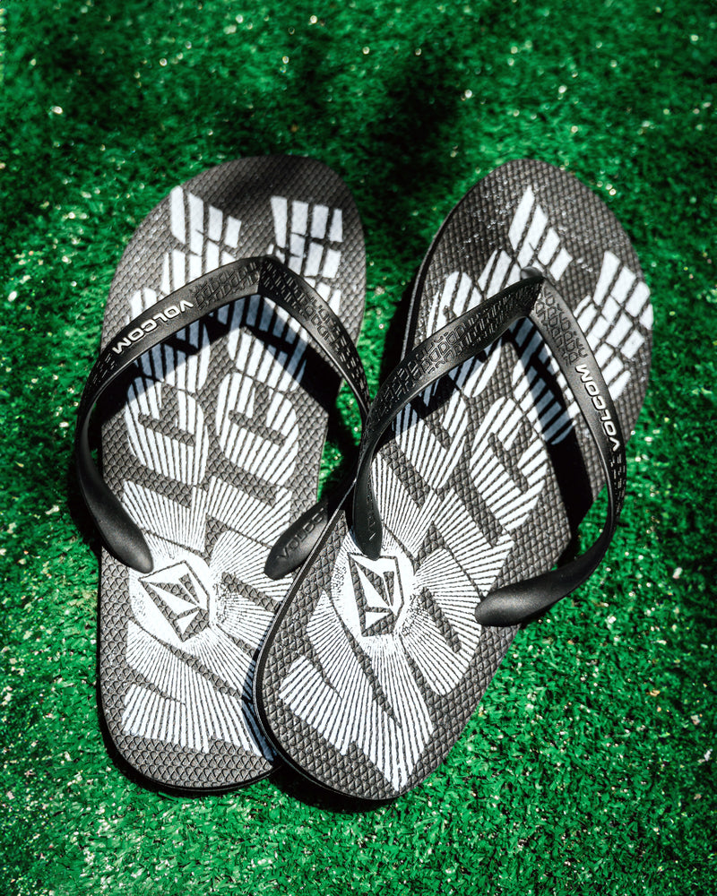 Men's Volcomotion Sandals by Volcom, featuring a bold text design in black and white, rest on vibrant green artificial grass under sunlight, delivering everyday comfort and signature style to your summer sandal rotation.