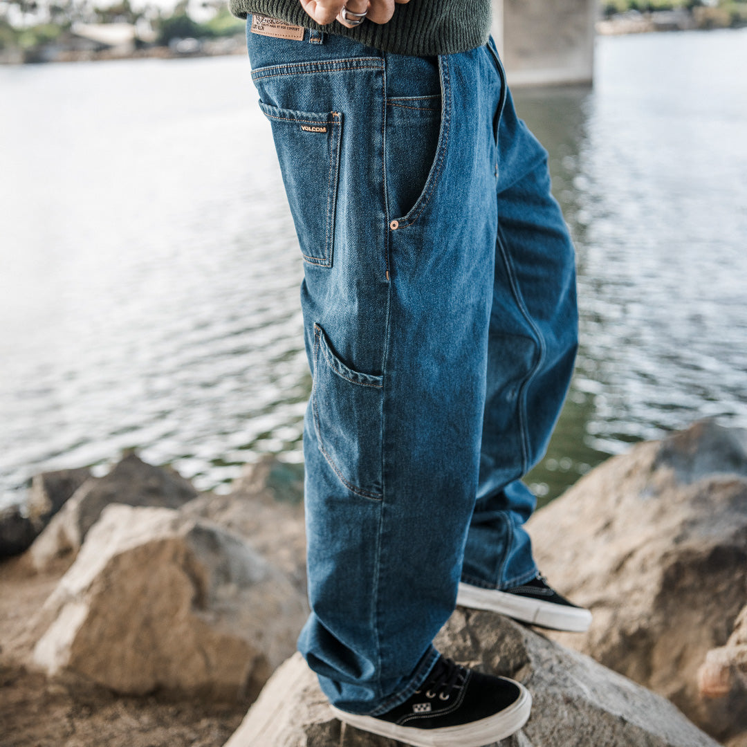 A person wearing loose-fit blue denim jeans with cargo pockets, standing on rocks near a body of water. The jeans are shown from the waist down, paired with black sneakers.