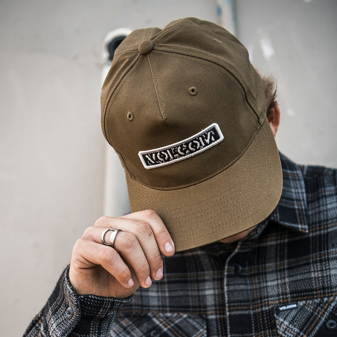 A person wearing a brown baseball cap with a white embroidered patch that reads 'VIKCIN' in white letters, holding the brim of the hat with one hand while wearing a dark plaid flannel shirt.