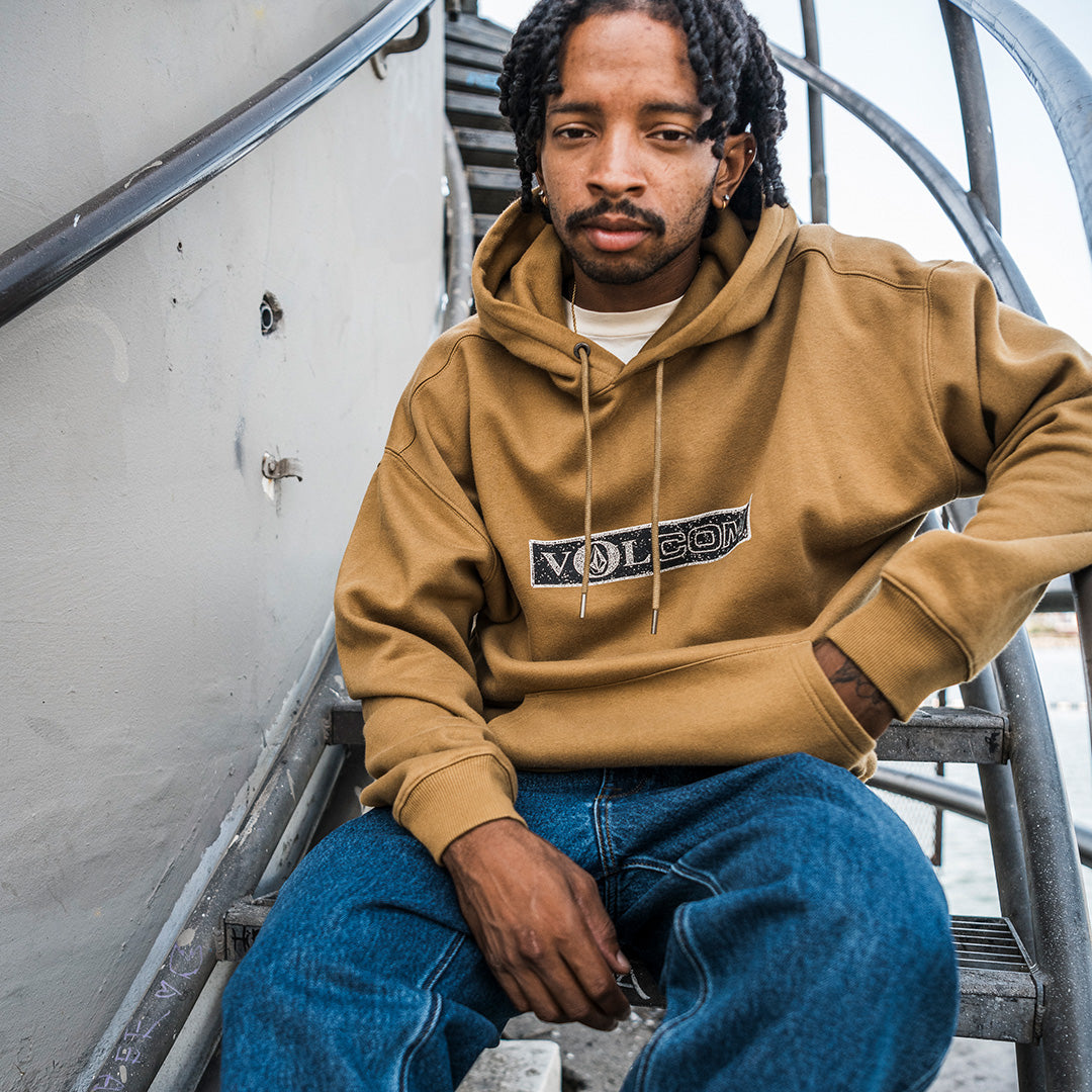 A man wearing a tan pullover hoodie with a 'Volcom' logo across the chest, paired with blue jeans, sitting on metal stairs.
