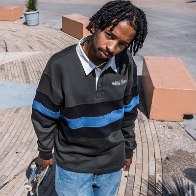 A man wearing a black and blue striped long-sleeve polo shirt with a white collar, standing outdoors on a paved area and holding a skateboard.