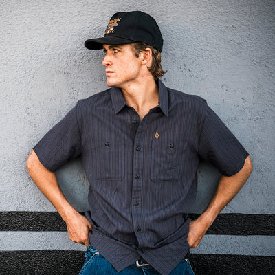 A man wearing a dark gray short-sleeve button-up shirt with a small embroidered logo on the chest pocket, paired with a black baseball cap and blue jeans, leaning against a gray textured wall.