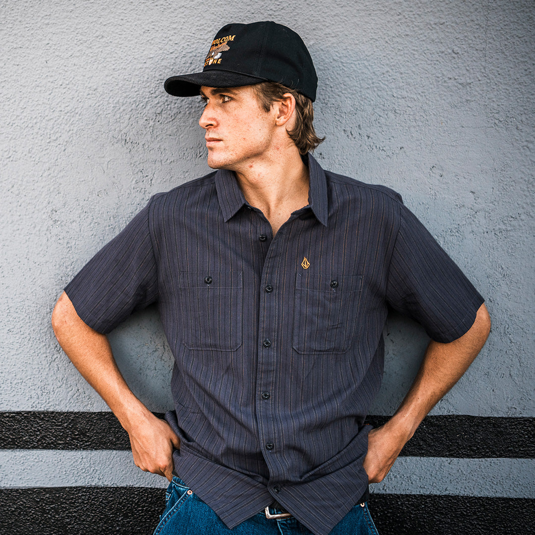 A man wearing a dark gray short-sleeve button-up shirt with a small embroidered logo on the chest pocket, paired with a black baseball cap and blue jeans, leaning against a gray textured wall.