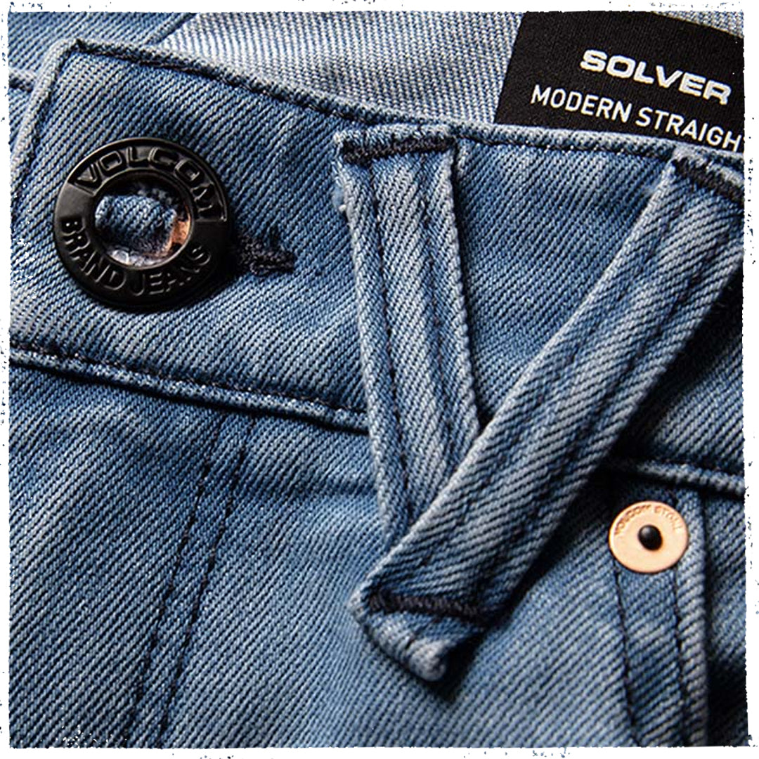 Close-up of blue denim jeans showing the waistband, button closure, belt loop, and a black brand label reading SOLVER MODERN STRAIGHT.