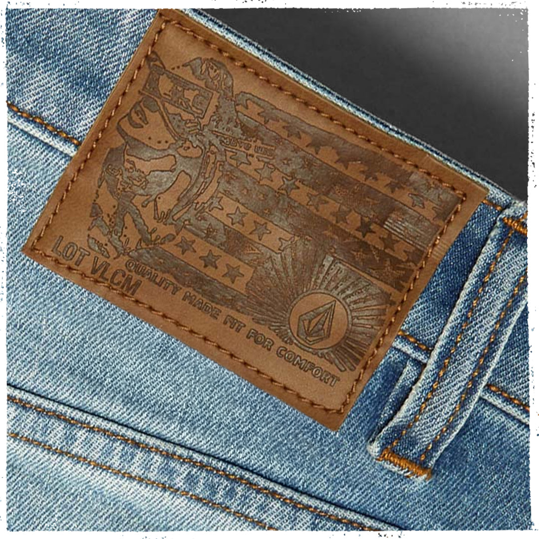 Close-up of a light blue denim jeans waistband showing a brown leather patch with an embossed American flag design and the Volcom mountain logo.