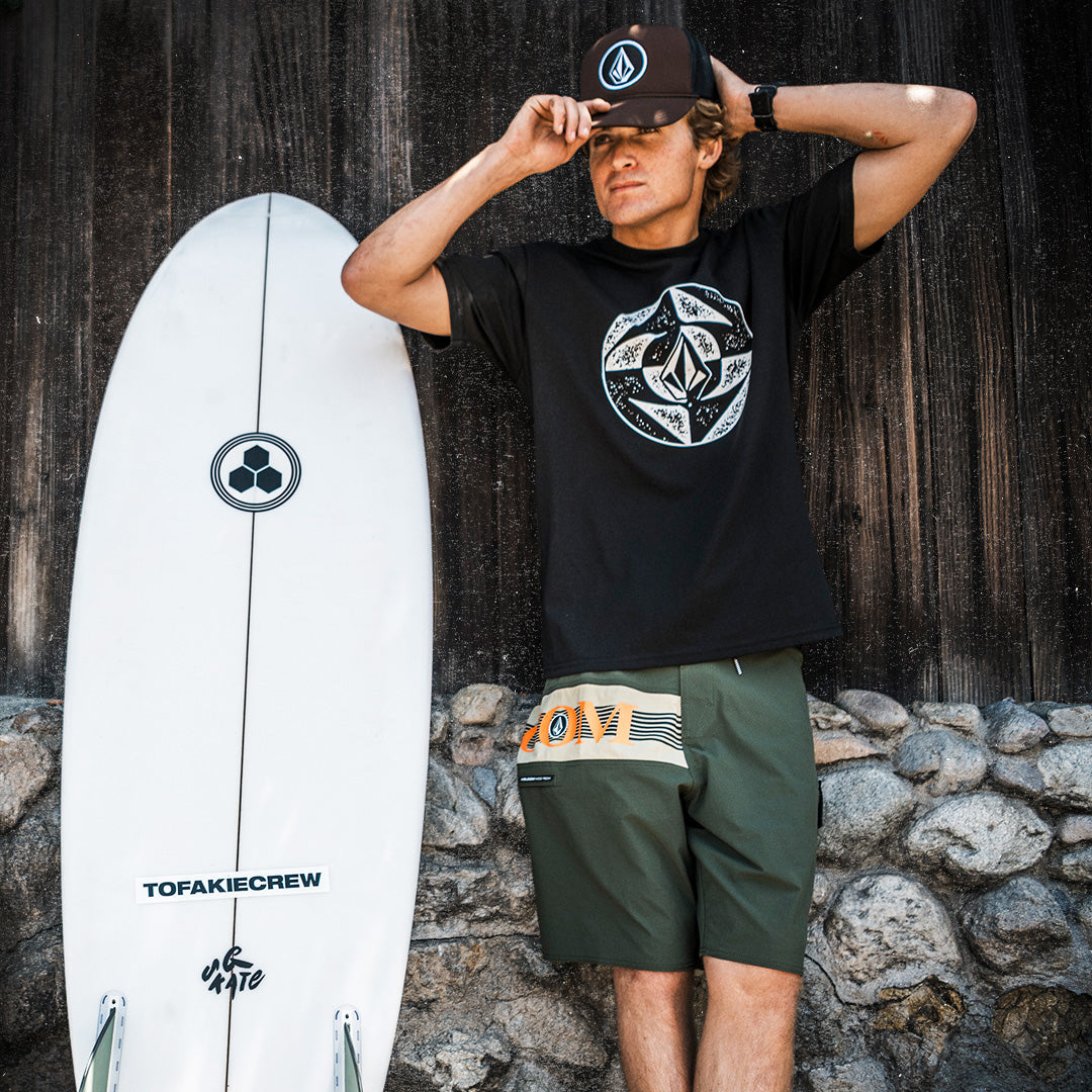Person wearing black shirt and green shorts standing next to a white surfboard outside