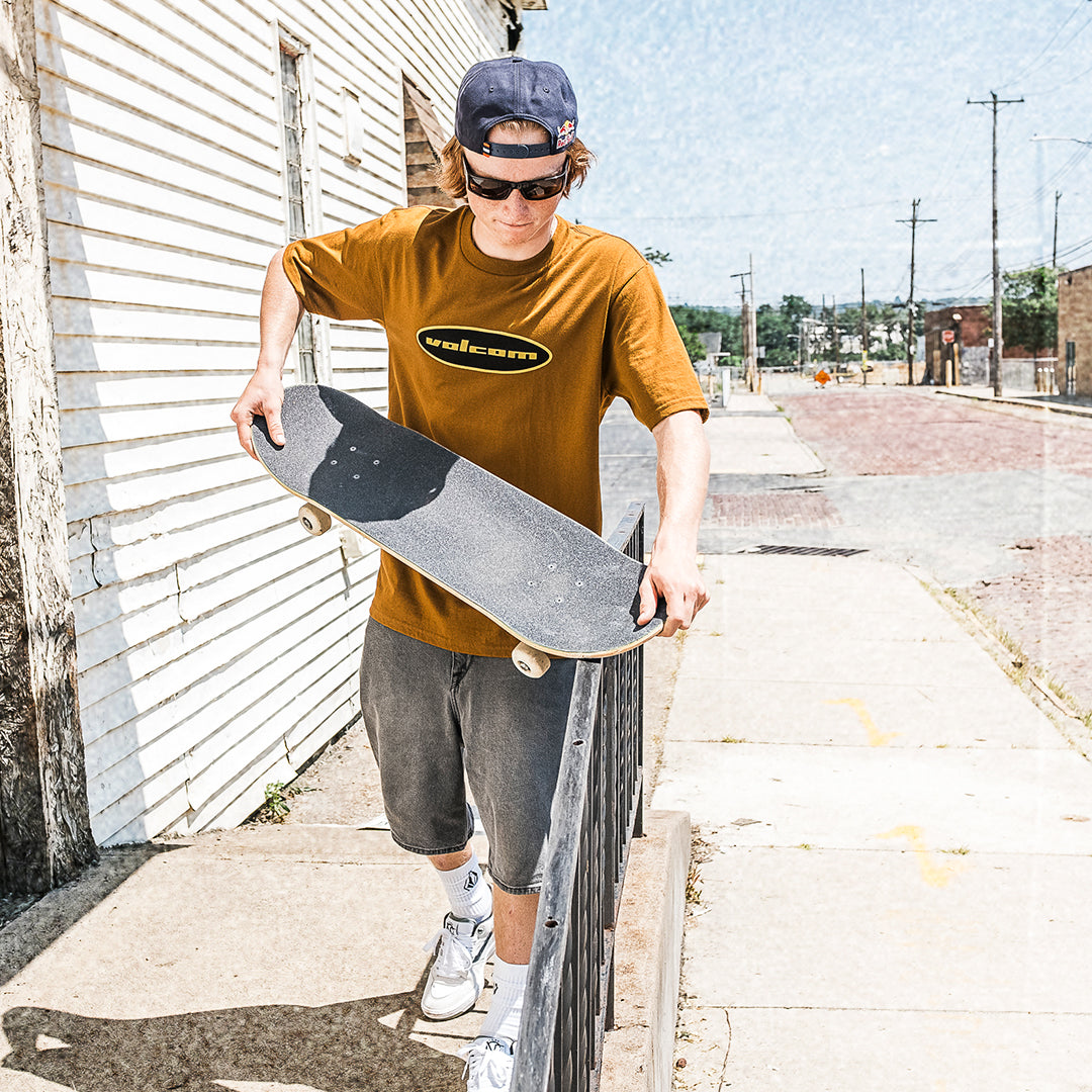 A young man wearing a brown t-shirt with a black and yellow 'Wicked' logo, gray shorts, and a navy cap holds a skateboard while standing on a sidewalk next to a white corrugated metal building.