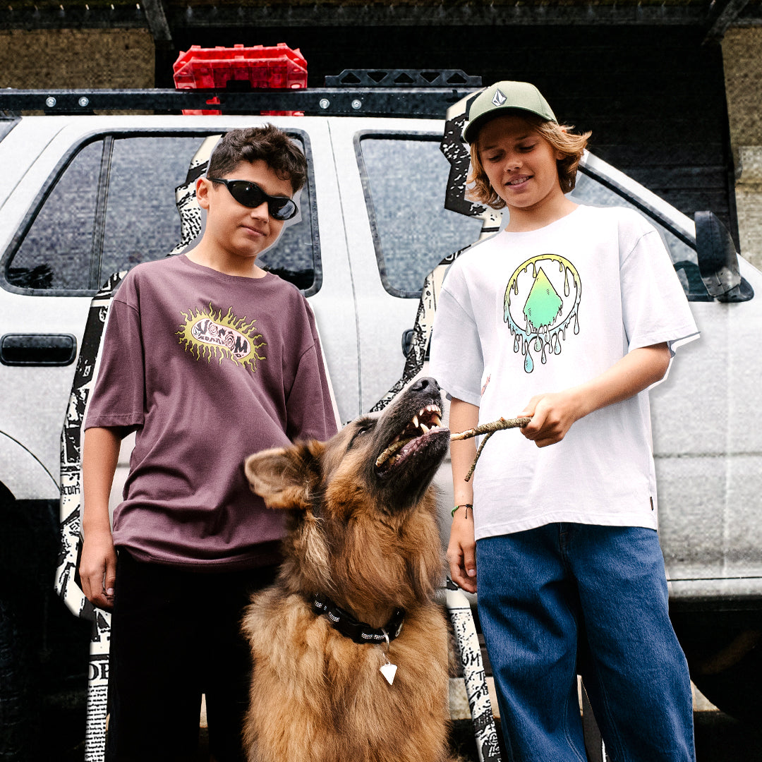 Two boys standing outdoors in front of a white pickup truck with a dog on a leash. The boy on the left wears a mauve-purple graphic t-shirt and black pants, while the boy on the right wears a white graphic t-shirt, blue jeans, and a green cap.