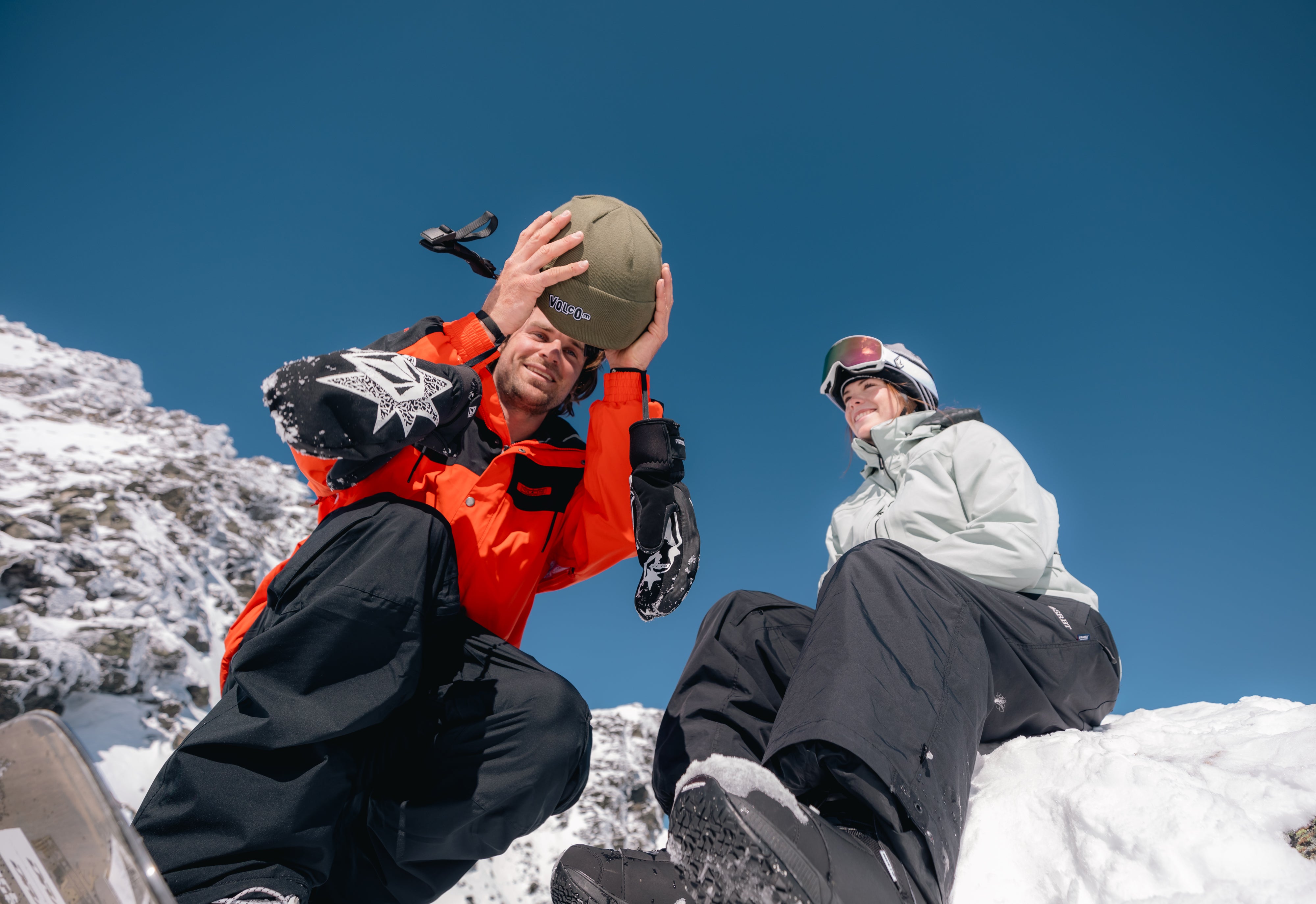Two people dressed in winter gear sit on snowy rocks under a clear blue sky. One person in an orange jacket adjusts a hat, while the other in white and black attire smiles, wearing ski goggles. Snowy mountains are visible around them.
