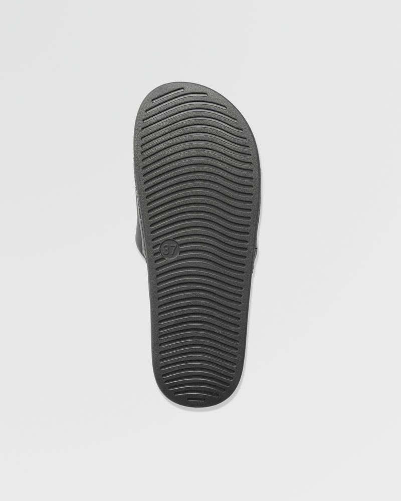 The image displays the black sole of Volcom Women's Volcation Slides, featuring horizontal grooves for traction and a size 37 mark. Set against a plain white background, it highlights everyday comfort and signature Volcom style.