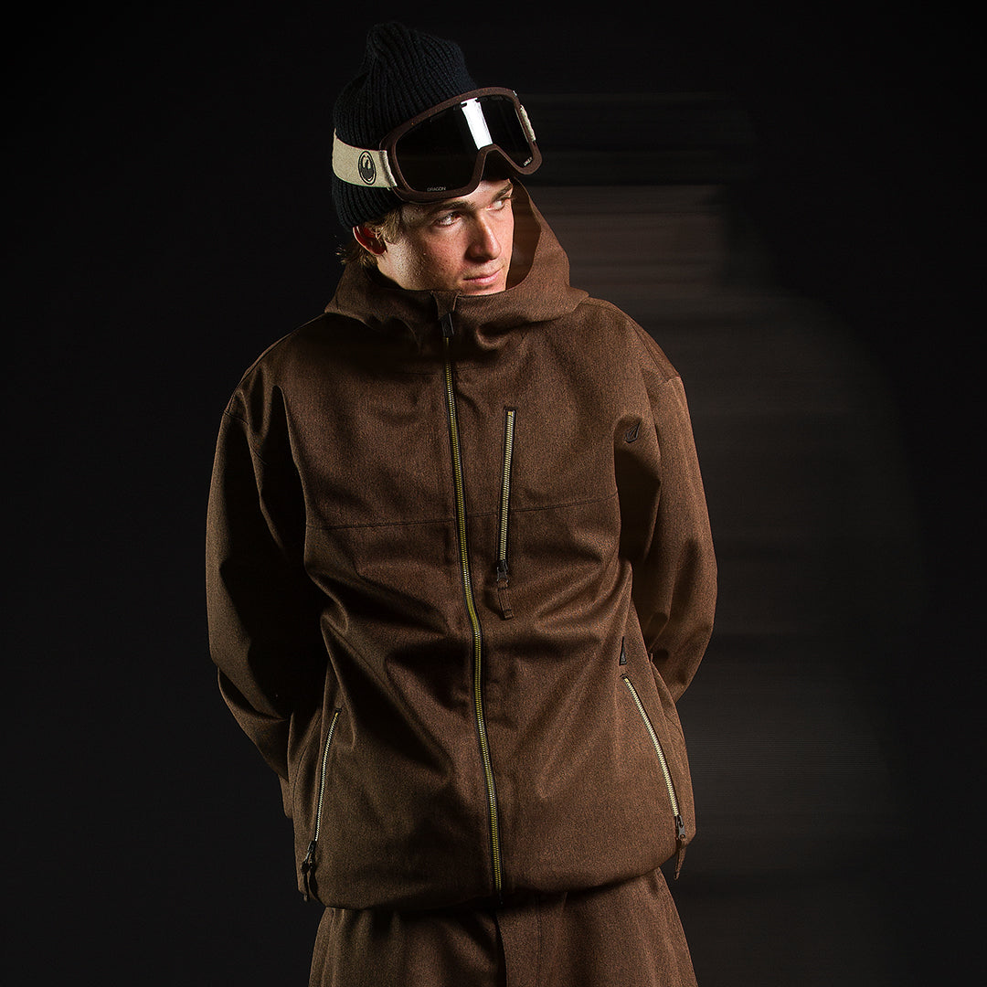 A man wearing a brown hooded snowboard jacket, matching brown pants, a dark beanie, and ski goggles, posed against a dark studio background.