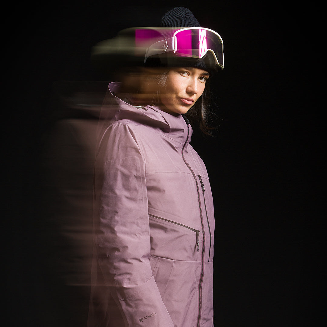 A woman in a light purple ski jacket and dark beanie with pink-tinted ski goggles, captured in a motion-blurry studio shot against a black background.