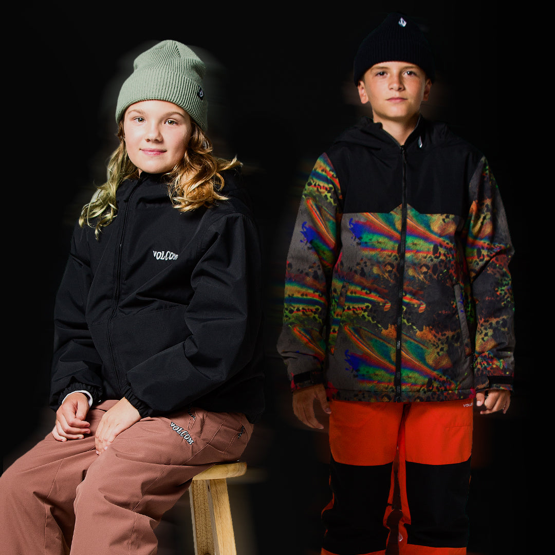 Two children modeling winter sports apparel against a black studio background. The child on the left wears a black Volcom jacket, a light green beanie, and brown pants, sitting on a wooden stool.