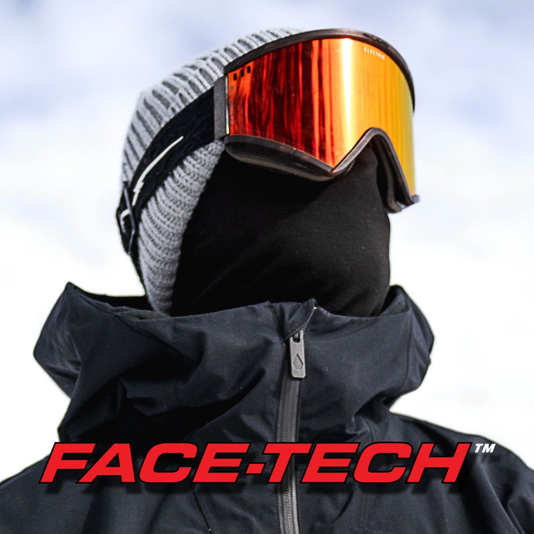 Close-up of a person wearing ski goggles with a reflective orange-red lens, a gray knit beanie, a black face mask, and a black jacket against a pale sky background. Large red 'FACE-TECH' text appears at the bottom.
