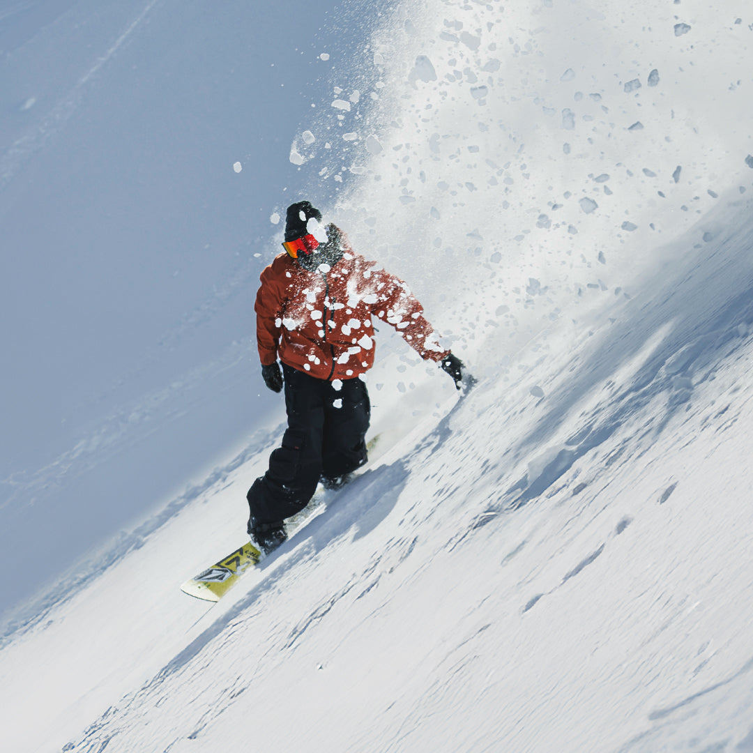 A snowboarder in a rust-orange jacket and black pants carving through fresh powder on a snowy mountain slope, with snow spraying up behind them.