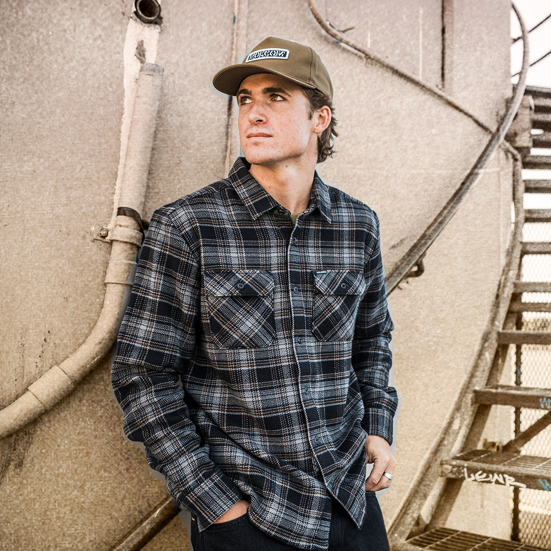 A man wearing a dark navy and gray plaid button-down flannel shirt and a tan baseball cap with a 'VOLCOM' patch, leaning against a textured concrete wall next to an outdoor metal staircase.