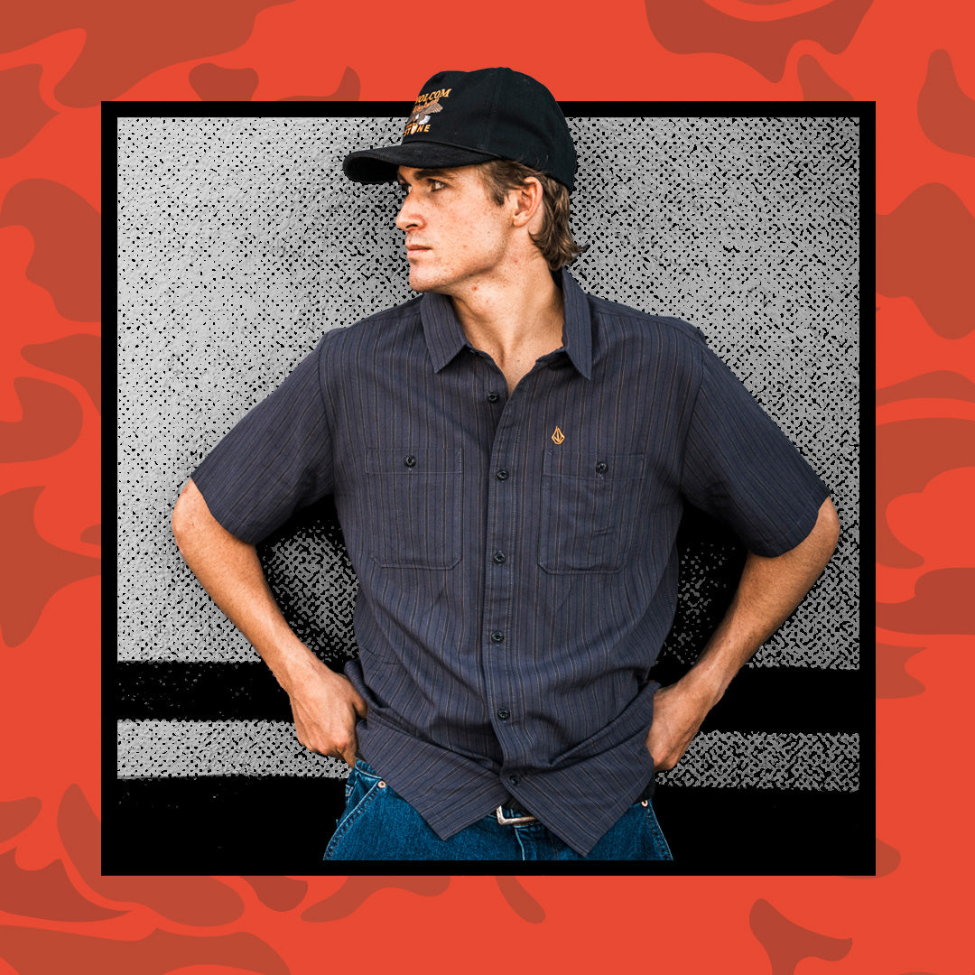 A man wearing a dark navy short-sleeve button-up shirt with thin vertical pinstripes, blue jeans, and a black baseball cap, posed against a textured gray background with a red camouflage border.
