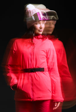 A model wearing a bright red ski jacket with a black belt, matching red ski pants, and a knit beanie with ski goggles, set against a dark background with a motion-blurred duplicate figure behind her.