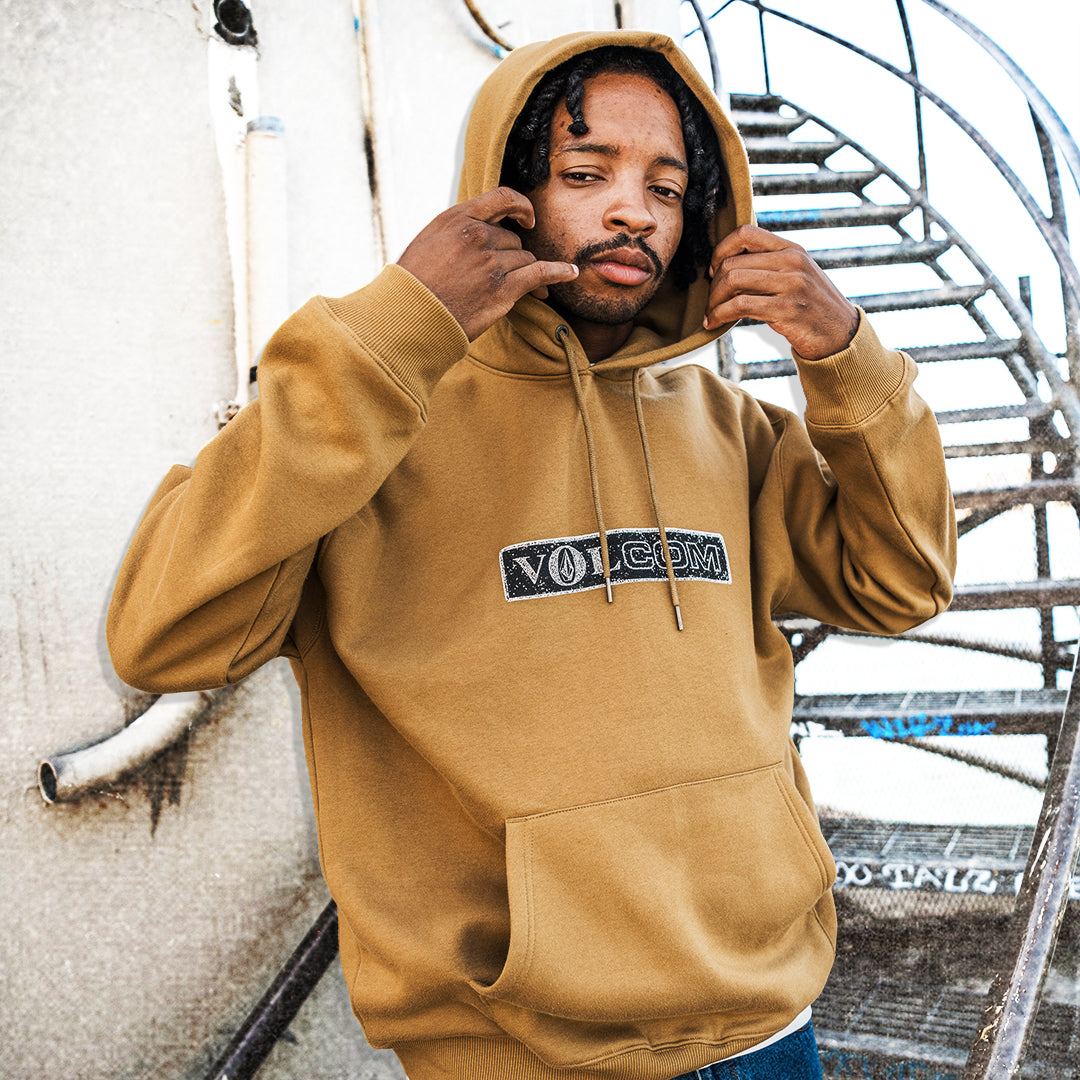 A man wearing a tan Volcom pullover hoodie with a black and white logo on the chest, posing in front of an outdoor metal spiral staircase.