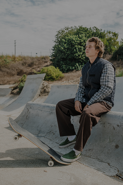 Person in a blue vest sitting on a skate ramp with a skateboard outside.