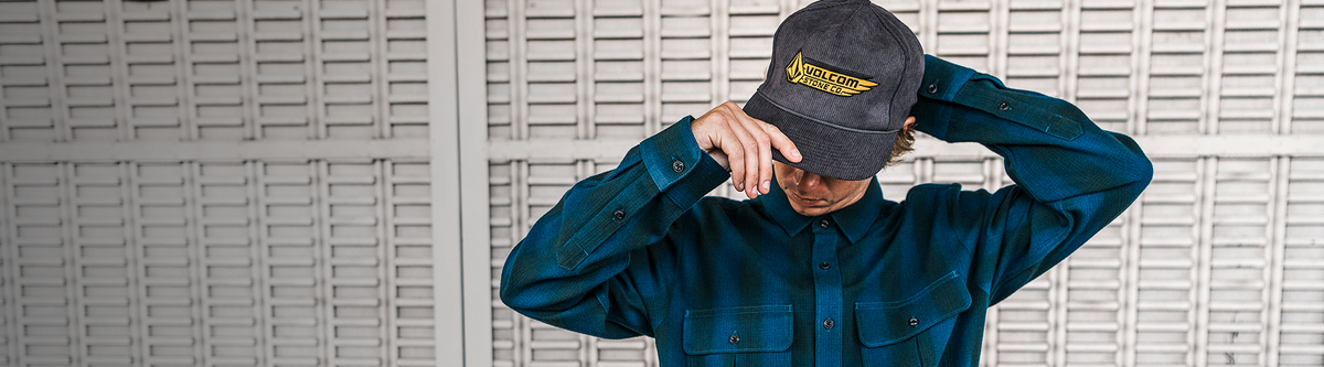 A person wearing a dark gray baseball cap with a yellow embroidered logo and a teal button-up shirt, standing in front of a white slatted wall.