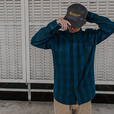 A person wearing a dark teal plaid button-up shirt, beige pants, and a dark gray baseball cap with a yellow logo, standing in front of a white slatted metal wall.