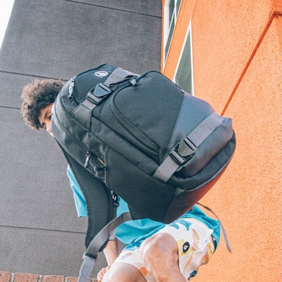 A person wearing a turquoise shirt and patterned shorts holds up a large black backpack, standing next to an orange building with a window, viewed from a low angle.