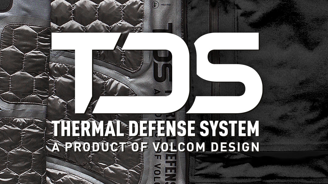 A close-up promotional graphic showing a black quilted jacket with large white text reading 'TDS' and 'THERMAL DEFENSE SYSTEM' plus 'A PRODUCT OF VOLCOM DESIGN.' The background is the jacket itself, and the visible brand is Volcom Design.